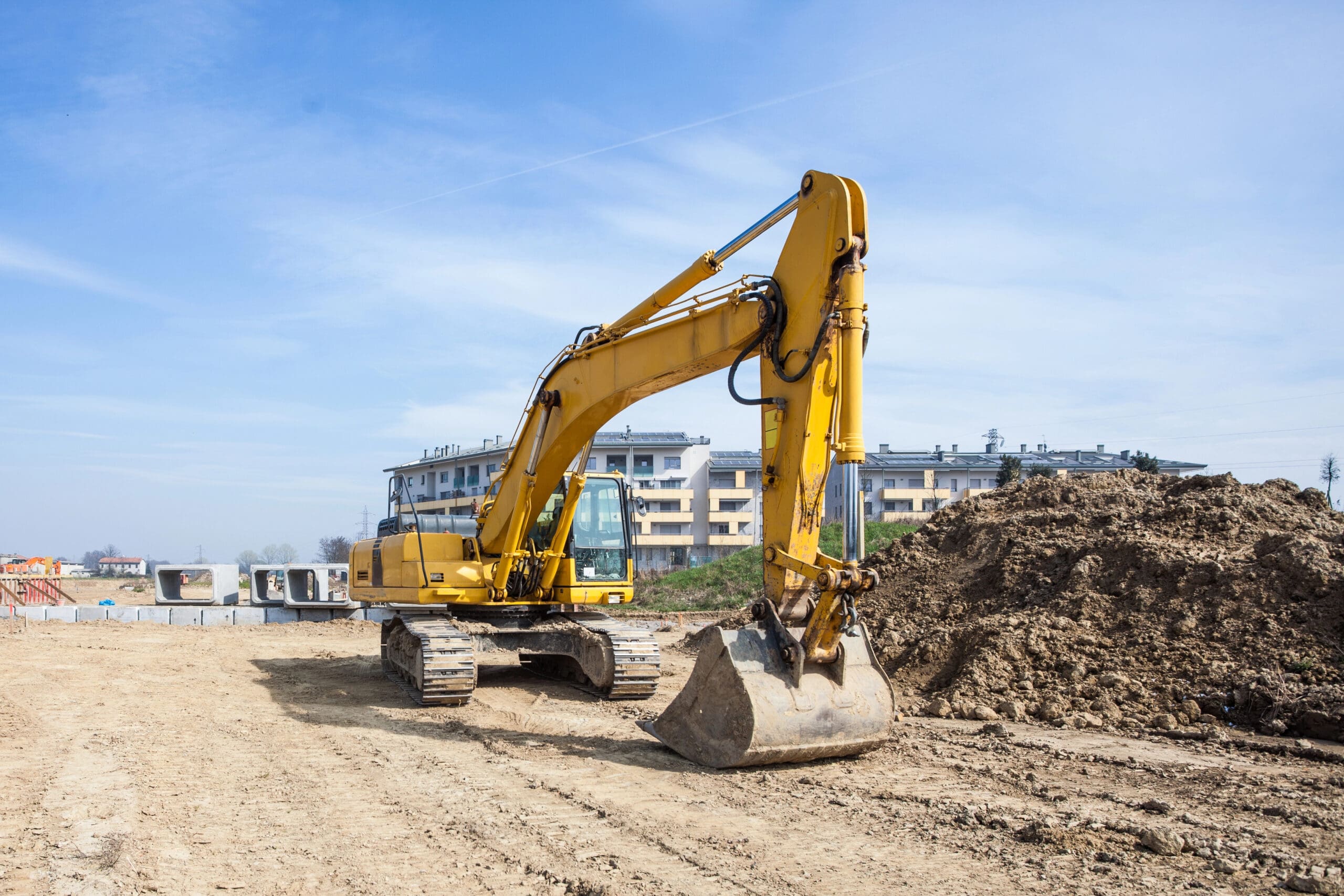 A yellow excavator with a large bucket is positioned on a construction site. The ground is uneven with dirt and debris, and there is a large pile of soil to the right. In the background, there are several residential buildings under clear blue sky.