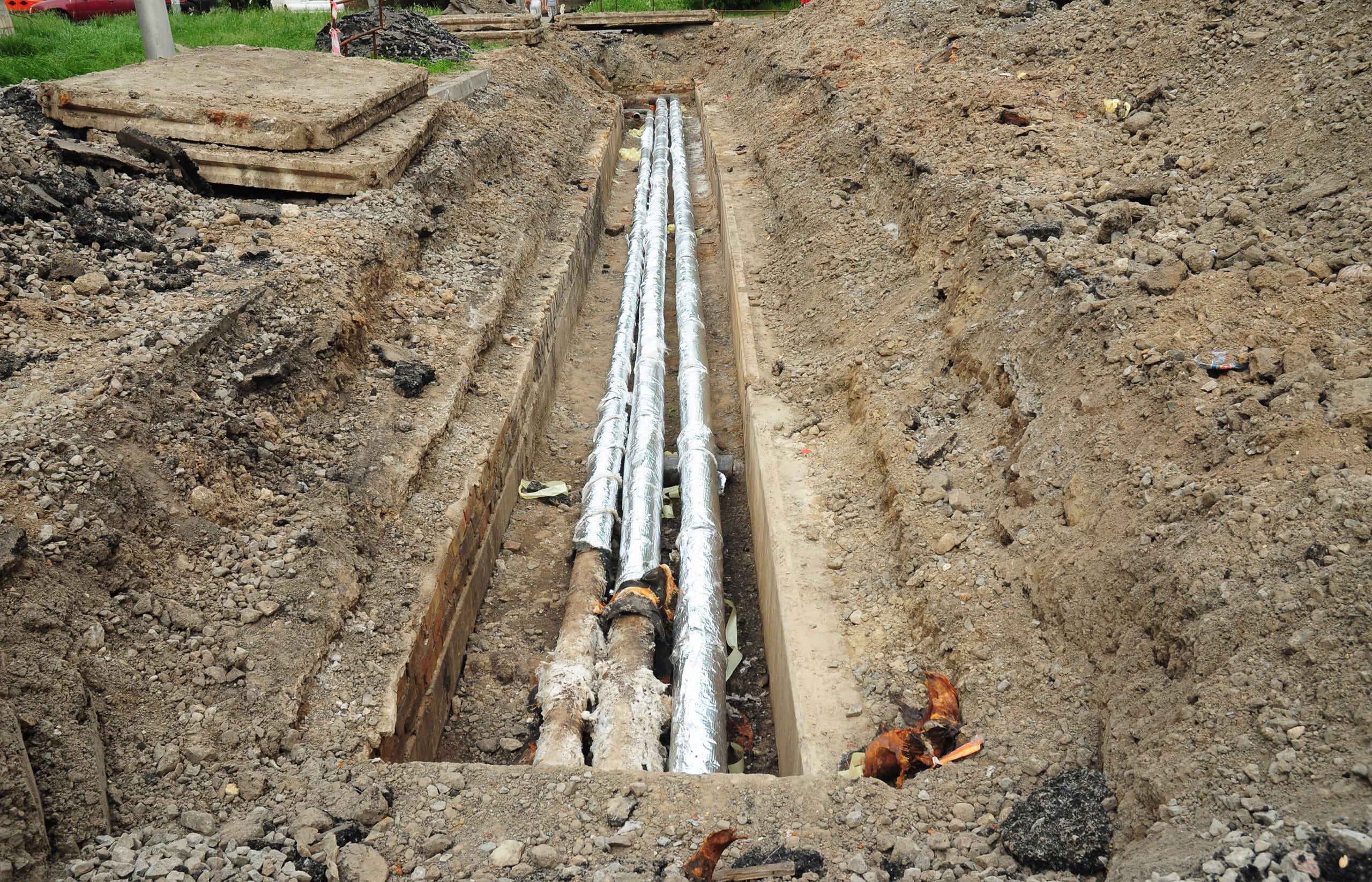 A trench dug in the ground with several large, insulated pipes laid side by side. The trench is surrounded by dirt and construction materials, suggesting ongoing infrastructure work.