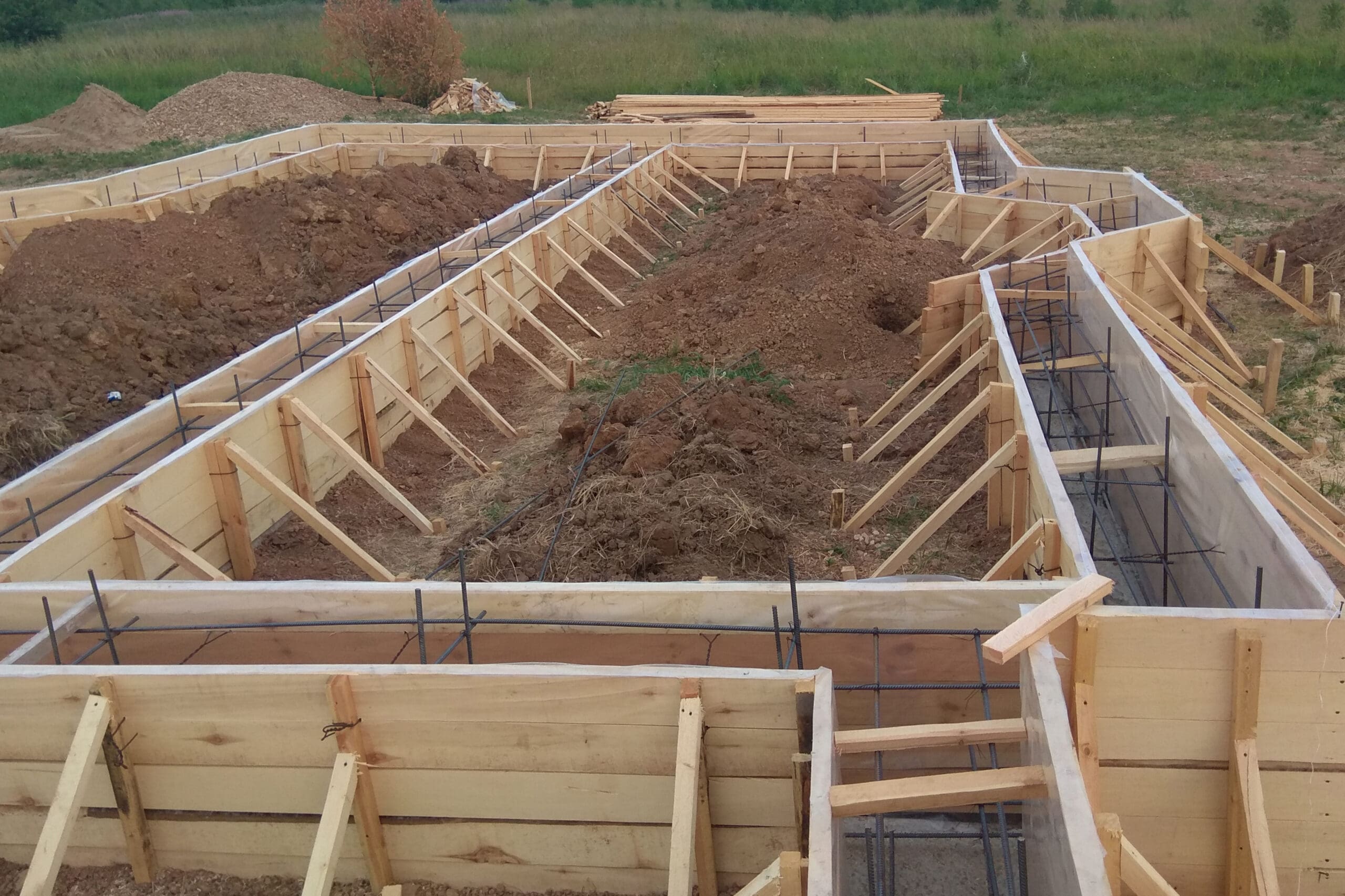 A construction site featuring wooden formwork for concrete foundations, with rebar grids in place. The formwork outlines several rectangular sections, prepared for pouring concrete. Piles of dirt and a grassy field are visible in the background.