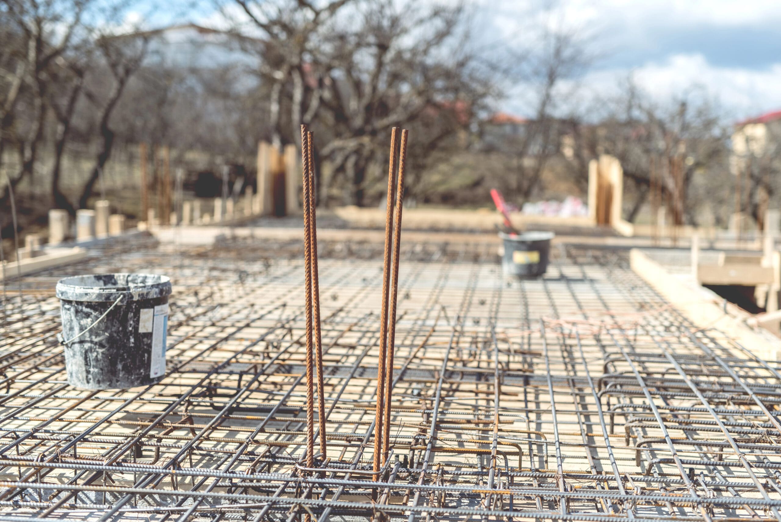 A construction site showing a concrete foundation in progress. Metal rebar and a grid of reinforcement structures are visible, with a gray bucket and a black bucket on the foundation. Trees and a partially cloudy sky are in the background.