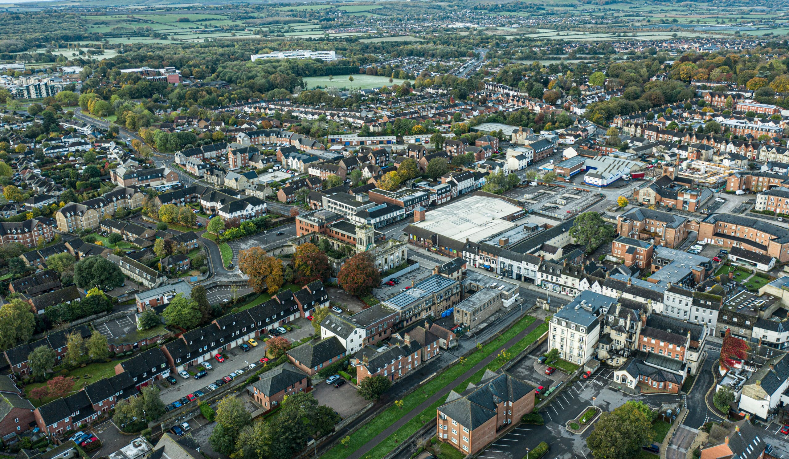 Aerial view of a densely built town with a mix of older and modern buildings, tree-lined streets, and green spaces. The landscape extends into the distance with fields and housing developments visible. Cars are parked along streets and in parking lots.