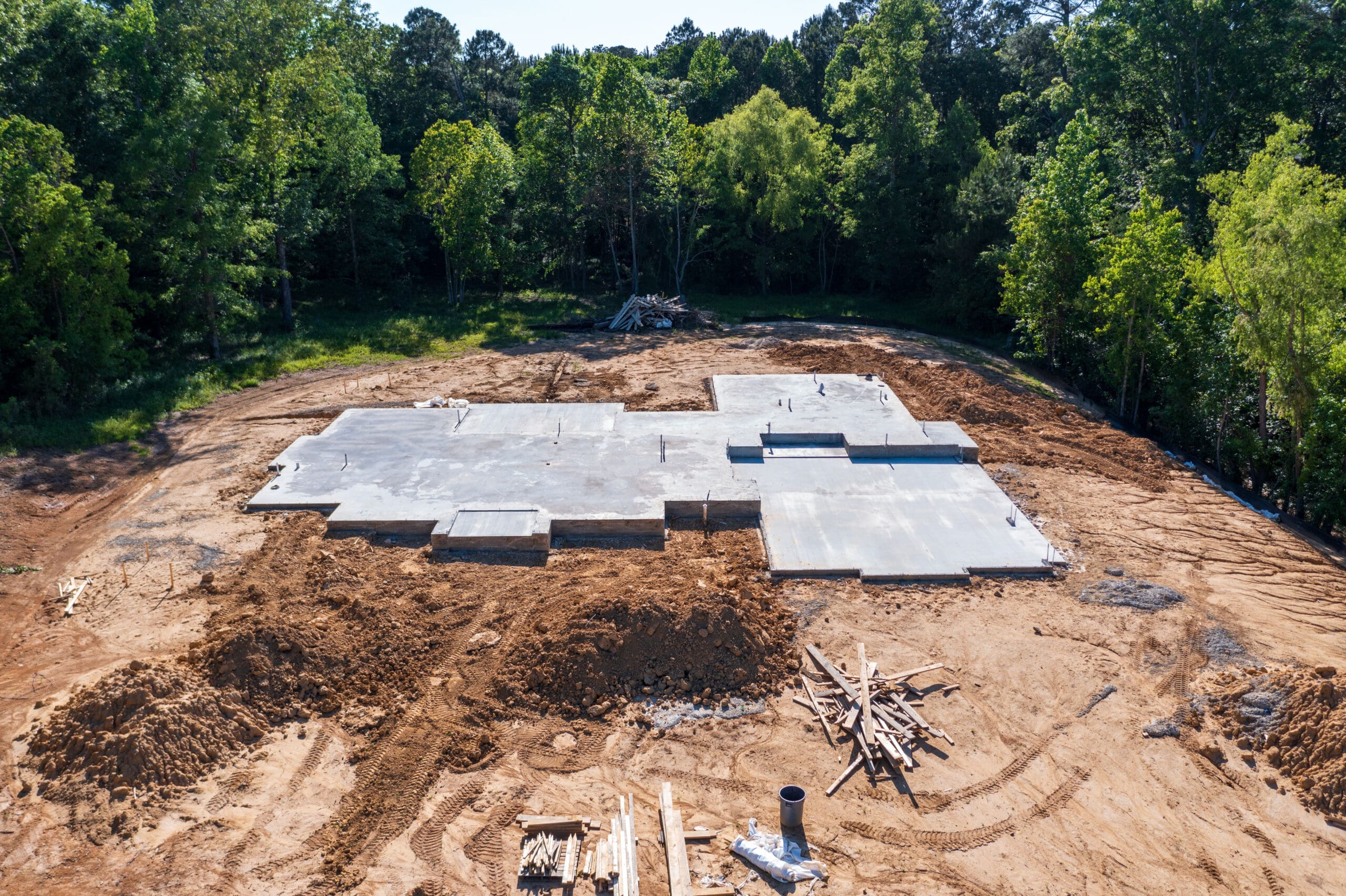 Aerial view of a construction site in a clearing surrounded by trees. The foundation of a structure is laid out, with wooden planks, pipes, and construction materials scattered around. Soil excavation is visible around the concrete foundation.