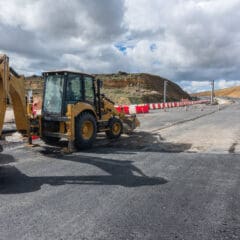 A yellow backhoe loader is parked on an asphalt-paved road under construction. The road is partially completed, with traffic cones and barriers lining the sides. The backdrop features rolling hills and a partly cloudy sky.