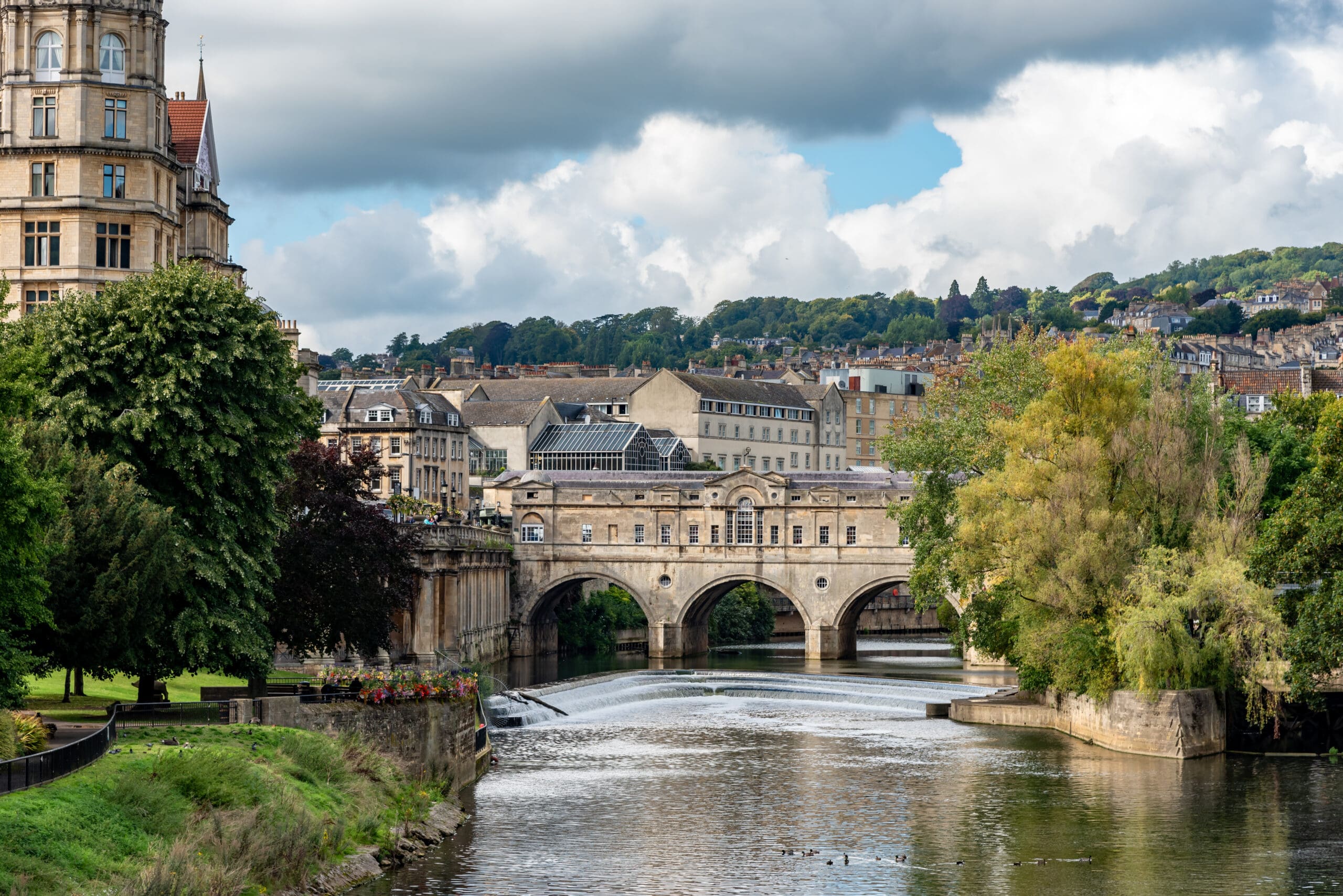 A scenic view of Pulteney Bridge in Bath, England, spanning the River Avon. The 18th-century bridge is adorned with shops and flanked by historic buildings and lush greenery. Ducks swim in the calm river, with hills and a cloudy sky in the background.