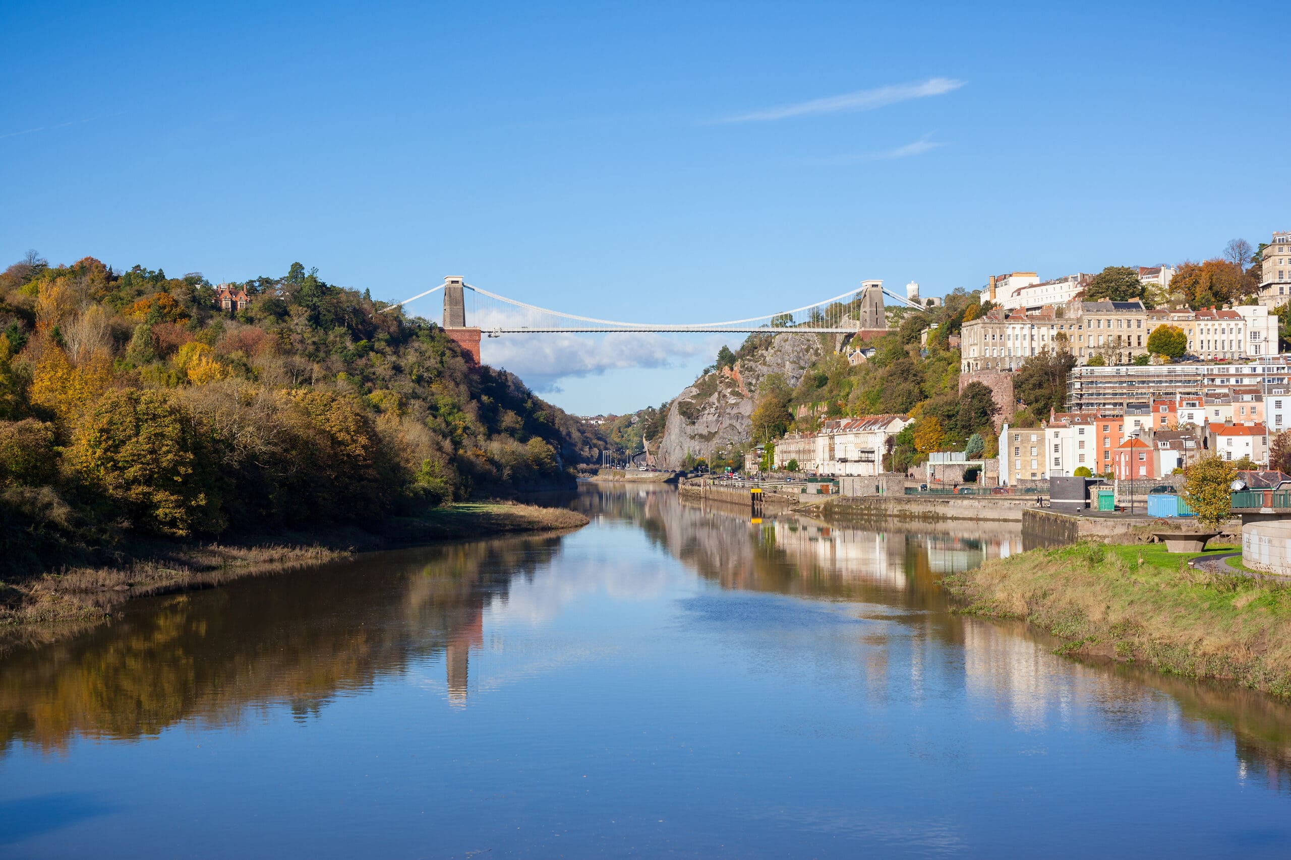 A scenic view of the Avon Gorge in Bristol, England, with the Clifton Suspension Bridge spanning across. The river flows calmly below, with lush green foliage and autumn-colored trees on either side, and buildings dotting the hillside under a bright blue sky.