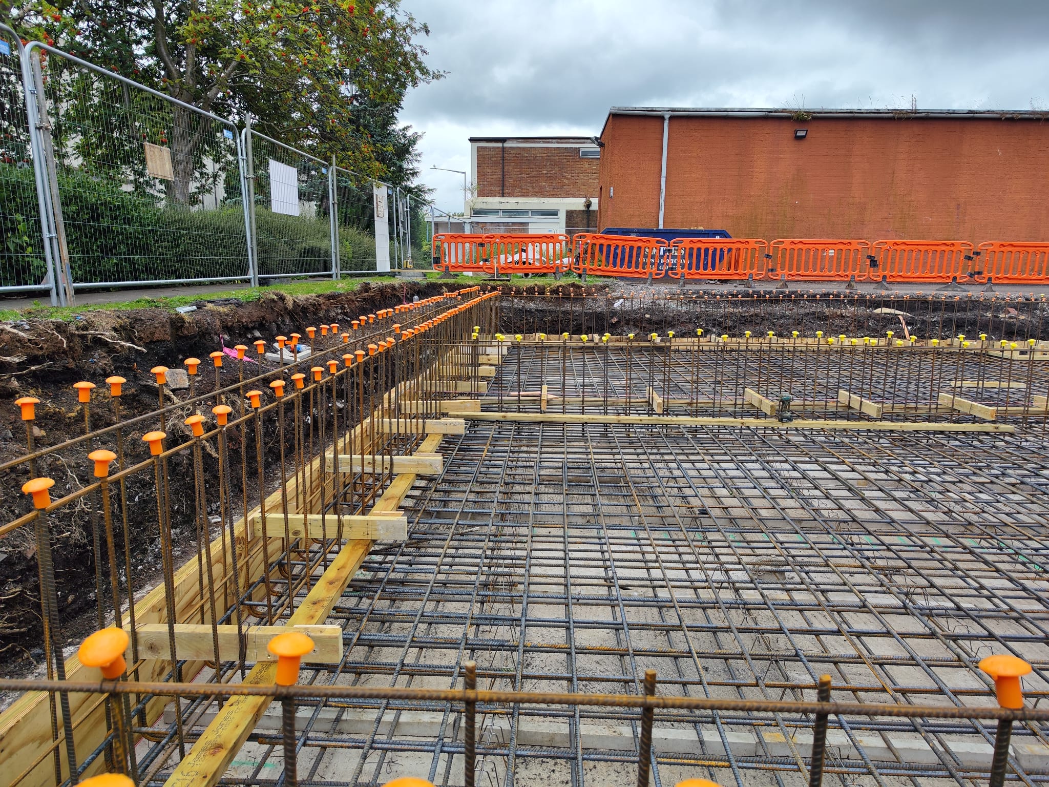 Construction site with steel reinforcement bars and yellow safety caps in place, preparing for concrete pouring. The rebar grid covers the ground, and wooden forms are set up. Safety barriers and a red building are in the background, with trees and a cloudy sky above.