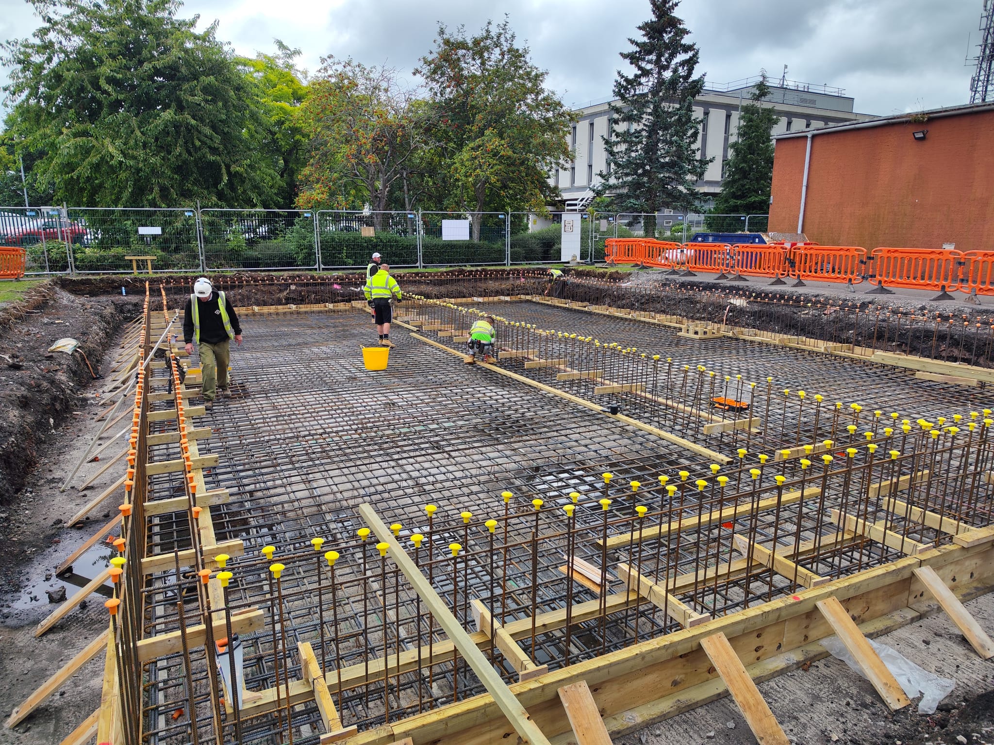 Construction workers are installing rebar in a large, rectangular foundation at a construction site. The area is surrounded by safety barriers and fencing, with trees and buildings visible in the background. The sky is overcast, indicating cloudy weather.