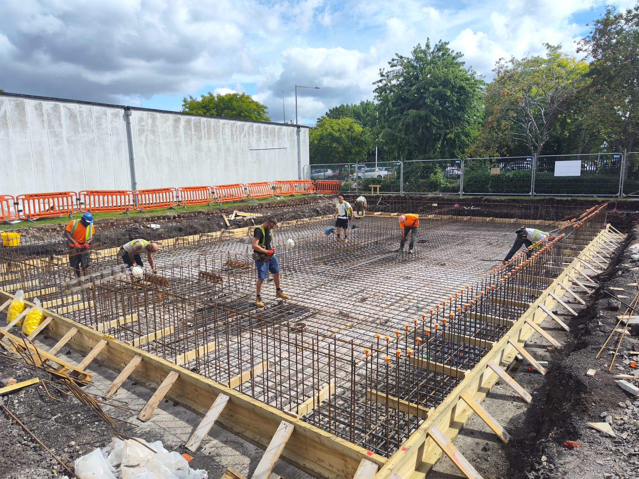 Construction workers wearing safety gear are working on the rebar foundation of a building site surrounded by wooden frames. Orange barriers and fencing are seen around the area. Trees and cloudy skies are in the background.