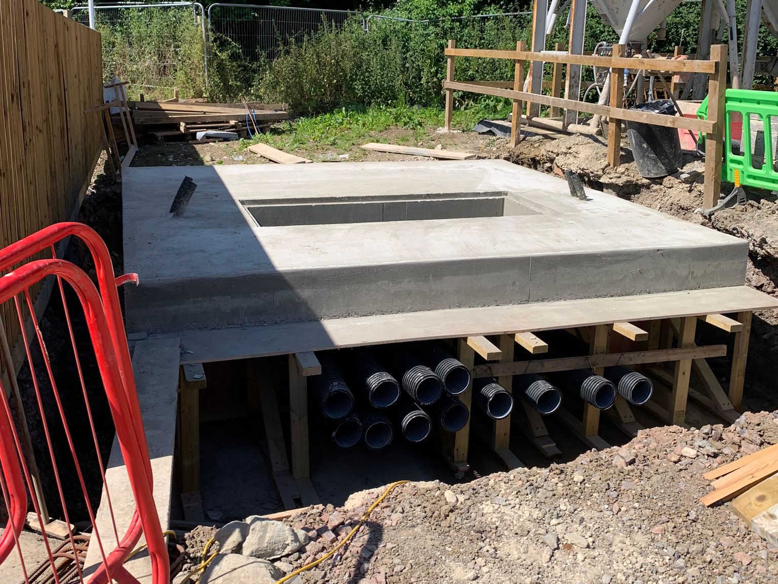 A construction site featuring a concrete foundation with an elevated central section. Multiple pipes extend under the structure, and wooden support frames are visible. Surrounding the area are fencing, barriers, and various construction materials.