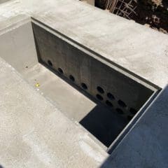 A newly constructed concrete drainage or utility trench, featuring a deep rectangular cavity with multiple circular holes in one of the trench walls. The surrounding area is a construction site with soil, rebar, and construction materials visible.