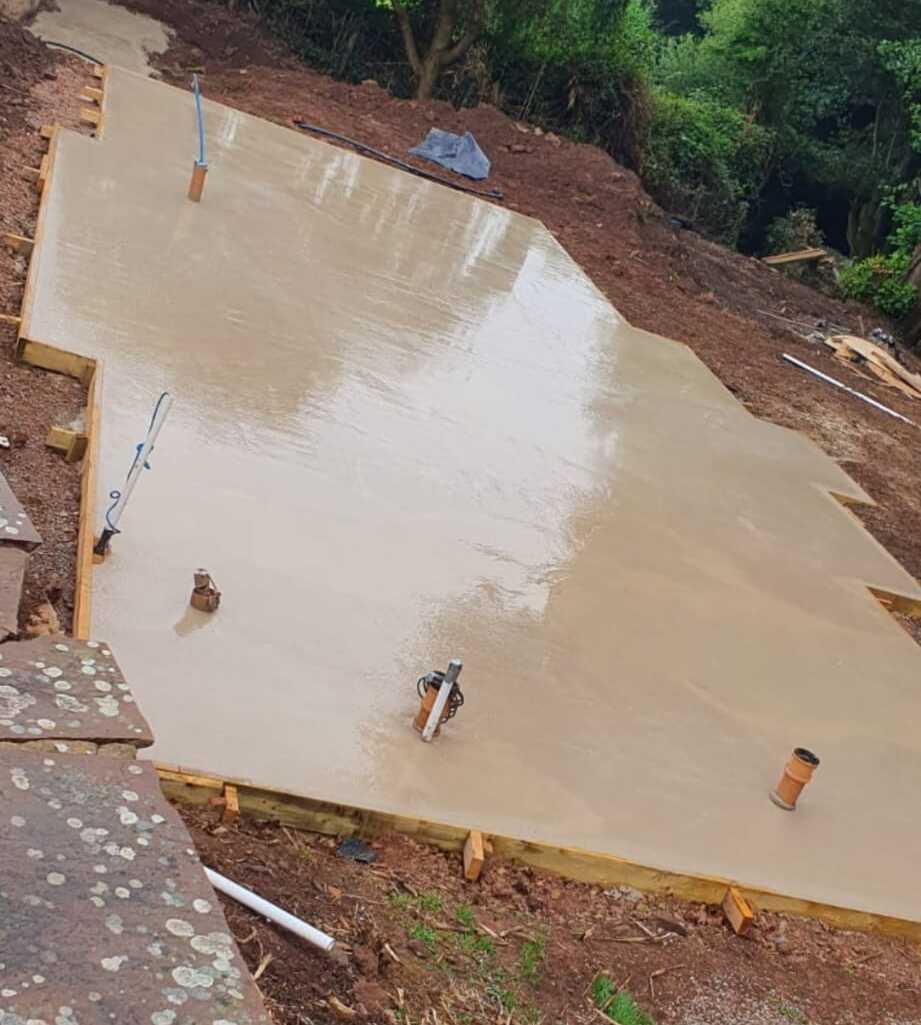 A newly poured concrete slab foundation with protruding pipes, set in a muddy and uneven construction site surrounded by green foliage. Some wooden framing is visible along the edges of the foundation.