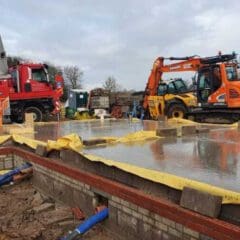 A construction site showing a large concrete foundation slab with visible concrete blocks and blue pipes. An orange excavator and a red crane truck are parked nearby. Workers in orange high-visibility clothing are present, and a portable restroom is visible in the background.