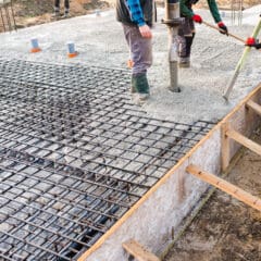 Construction workers are pouring concrete over steel reinforcement bars to create the foundation of a building. The workers are dressed in protective clothing and use tools to evenly distribute the concrete inside a wooden frame.