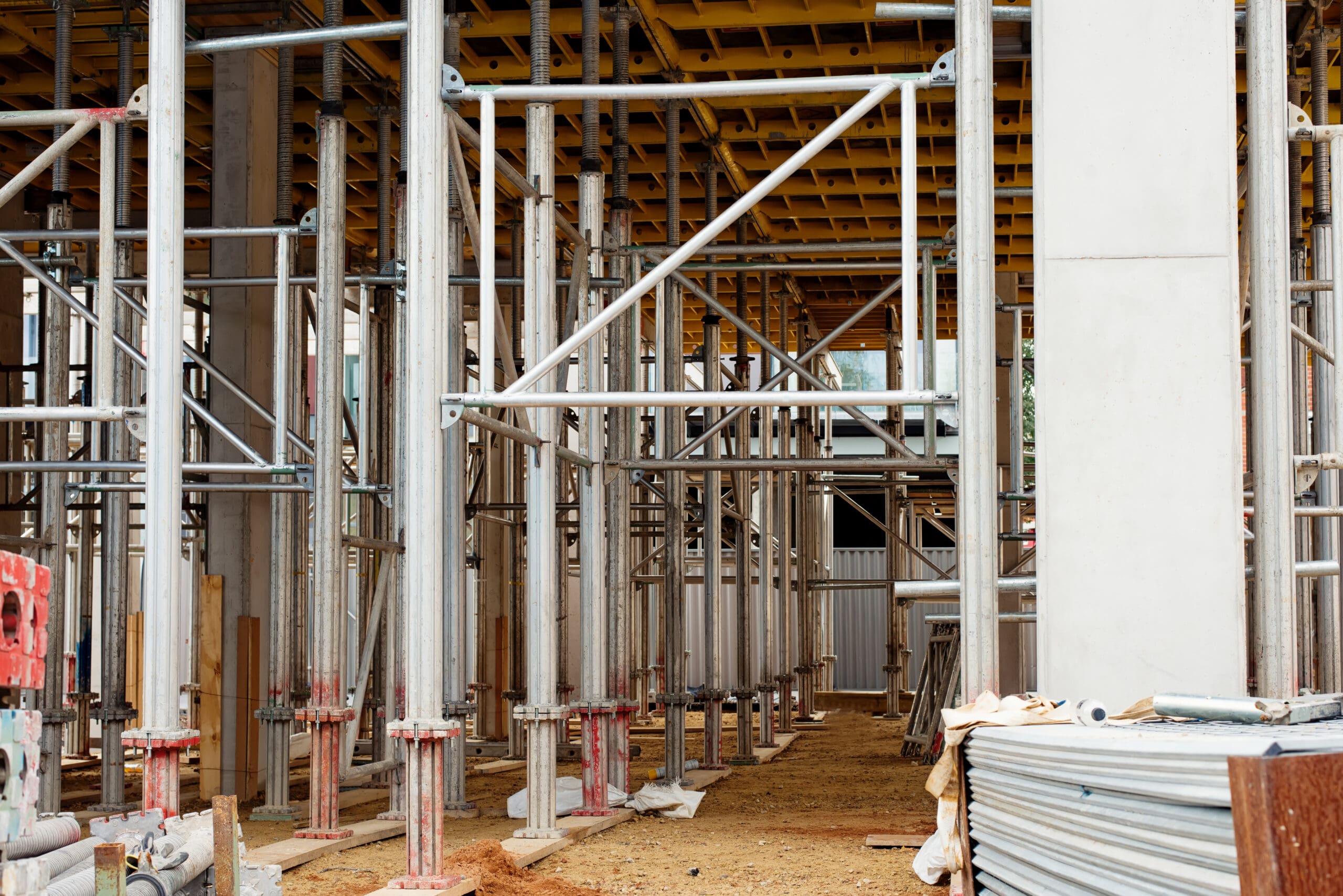 A construction site with multiple metal scaffolding structures and support beams arranged in a grid pattern. The ground is covered in dirt and construction materials. Sheets of metal and wooden boards are stacked on the right side. A partial ceiling is visible.