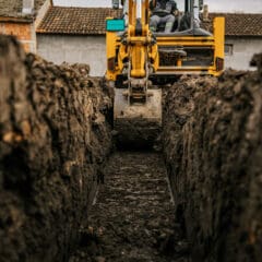 A yellow excavator operates in a deep, narrow trench, with large piles of soil on either side. The operator is partially visible in the cabin. In the background, there are buildings with tiled roofs. The scene is overcast, adding a muted tone to the environment.