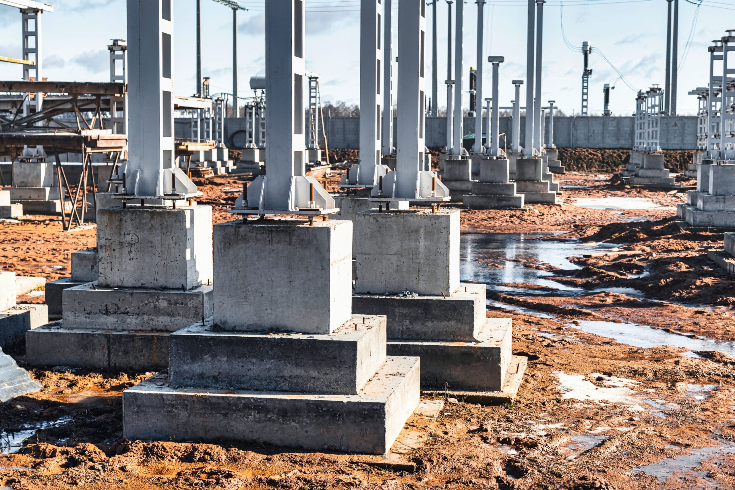 Construction site featuring multiple concrete support pillars, metal frameworks, and muddy ground. The pillars are set in a structured grid, secured in square concrete bases. Scaffolding and additional construction equipment are visible in the background.