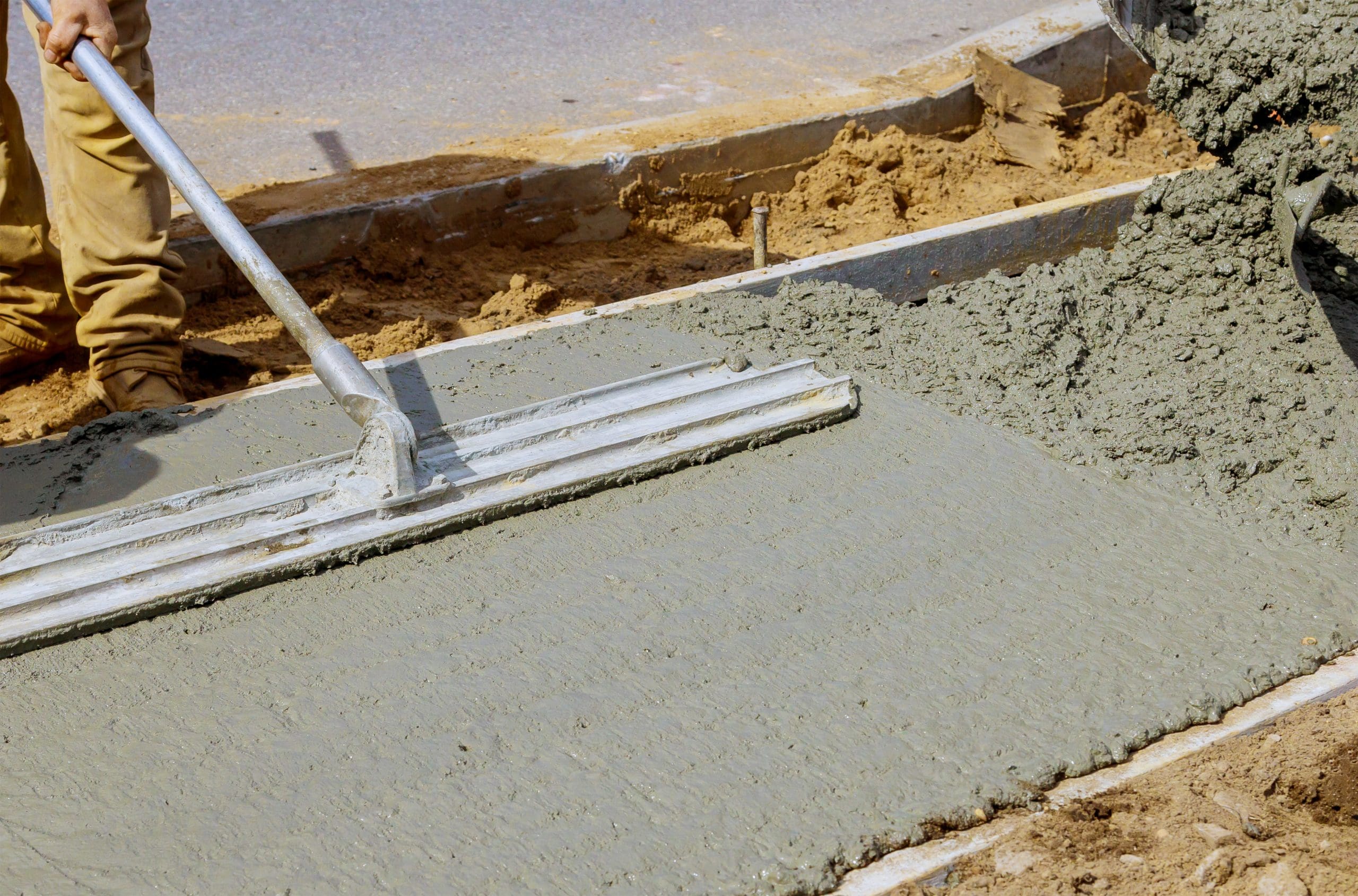 A construction worker smooths wet concrete with a large metal float tool at a building site. The surrounding area shows exposed soil and wooden forms, indicating ongoing construction work. The worker's legs and boots are visible in the scene.