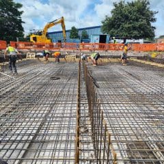 Construction workers are seen working on a building site, assembling a grid of steel rebar on a large concrete foundation. An excavator and safety barriers are visible in the background, along with a blue building and trees under a partly cloudy sky.