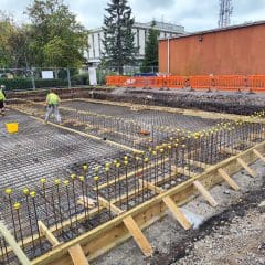 Construction workers are preparing a large foundation with numerous steel rebar grids, ready for concrete pouring. The construction area is bordered by a fence, and one side features an orange safety barrier. Trees and a building are visible in the background.