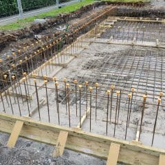 The image shows a construction site with rebar grids and wooden formwork set up for a concrete foundation. Wooden planks form the base frame, and rebar extends vertically and horizontally, connected with orange protective caps on top. A surveyor's tripod is visible on the left side.