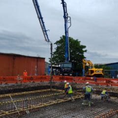 Construction workers in high-visibility vests work on a large concrete foundation, with a concrete pump truck in the background placing concrete into the framework. Orange safety barriers surround the construction site. A tree and industrial buildings are visible in the background.