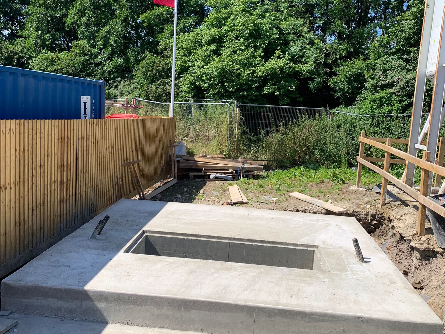 Construction site featuring a newly poured concrete foundation with a rectangular recess in the center. The area is surrounded by wooden fencing and containers, and there are building materials scattered around. In the background, there's dense greenery.