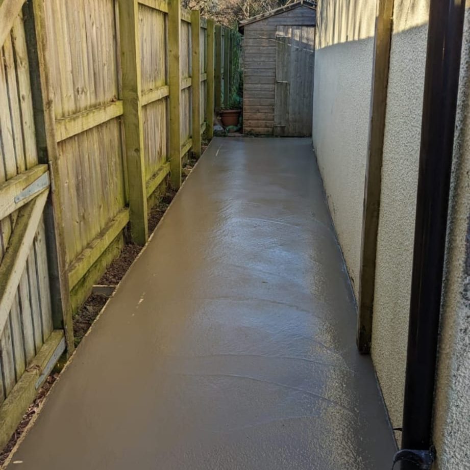 A narrow concrete pathway runs between a wooden fence on the left and a beige wall on the right. The pathway leads to a wooden shed at the end, with a couple of potted plants placed near the shed. The area appears clean and well-maintained.