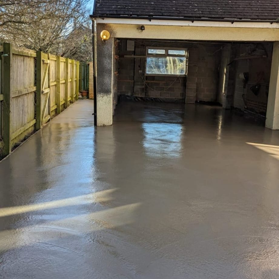 A freshly poured and smoothed concrete garage floor with a partially open garage door. The concrete is still wet and glossy, reflecting sunlight. A wooden fence runs alongside the driveway leading up to the garage. Trees and other houses are visible in the background.