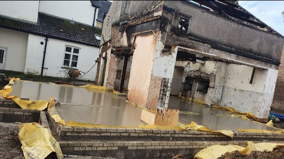 Partially demolished building with exposed interior walls, surrounded by a foundation filled with fresh concrete. Yellow tarps are draped nearby. The scene appears to be part of a construction or renovation project. Nearby buildings are intact.