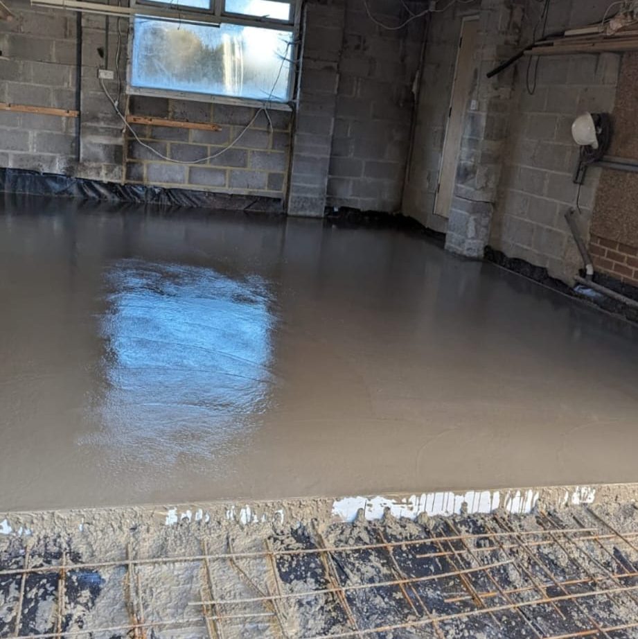 A room under construction with freshly poured concrete covering the floor. The walls are unfinished with exposed cinder blocks, insulation, and wiring. Sunlight is coming through a window in the back, reflecting off the wet concrete surface.