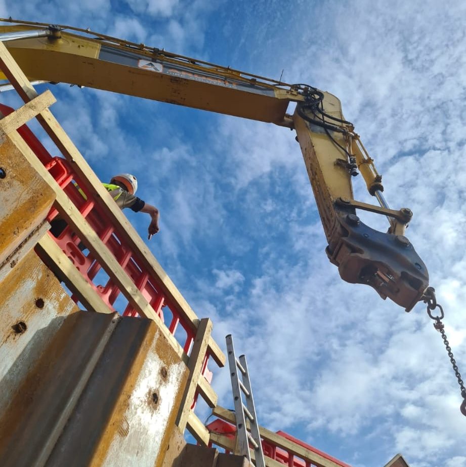 A construction worker in a hard hat and safety vest points downward while standing on a structure. A yellow excavator arm with a chain attached extends above the worker. The sky is partly cloudy in the background.