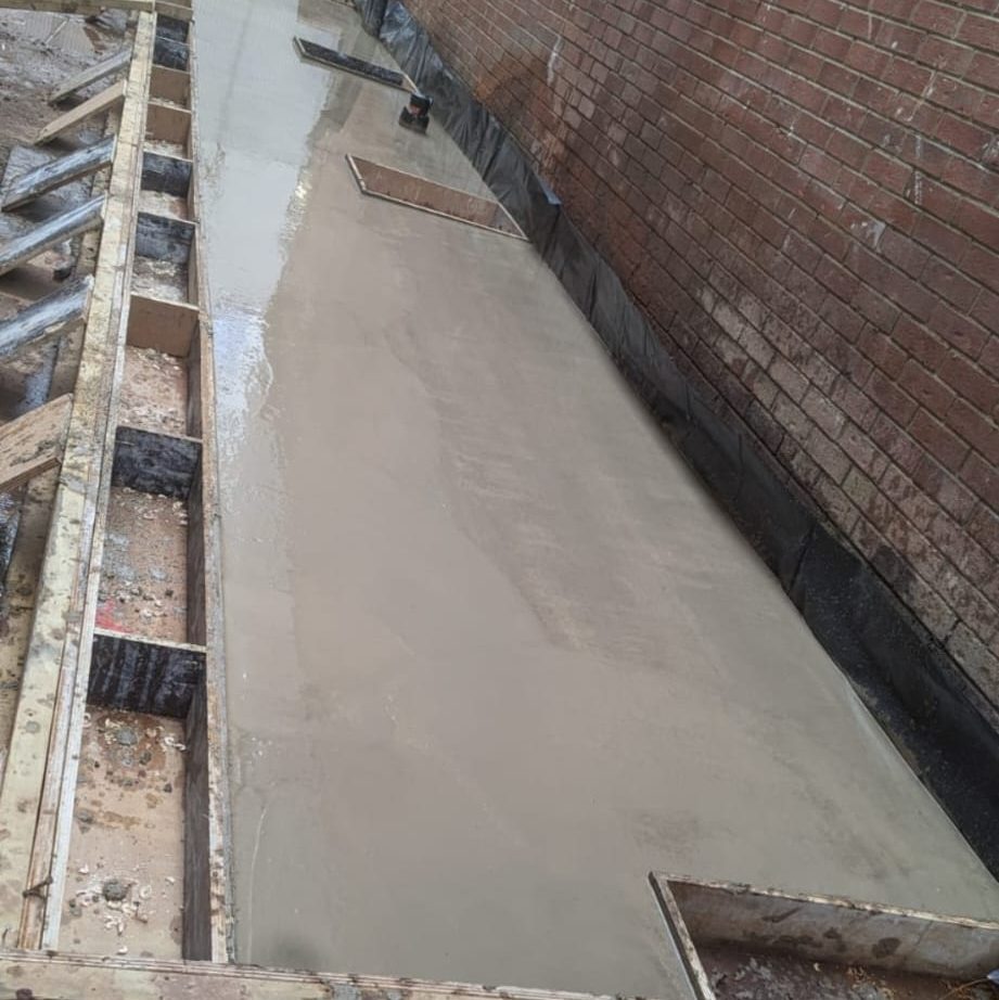 A freshly laid concrete pathway partially constructed against a brick wall. There are wooden frames and supports along the sides of the path, indicating ongoing construction. Some sections of the pathway are still wet and unfinished.