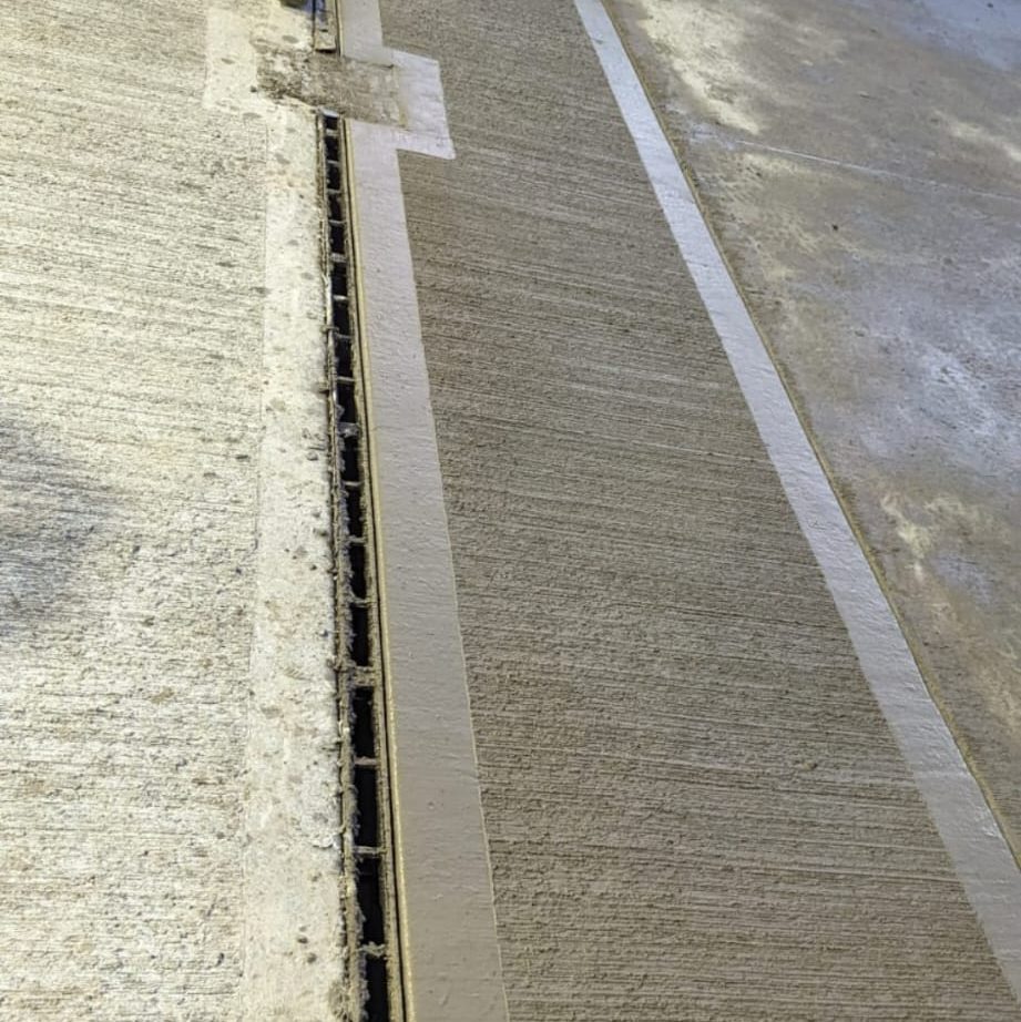 Concrete floor with a long, narrow textured strip running horizontally in the center. The strip includes a linear drainage system. The surrounding areas have smoother and rougher concrete finishes with some wear and discoloration visible.