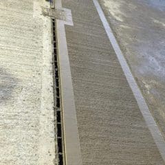 A worker in a safety outfit is spreading fresh concrete on a rectangular section of a floor, smoothing it evenly. Construction materials and tools are scattered around, including a trowel and a wooden board. Nearby, an unfinished and rough concrete edge is visible.