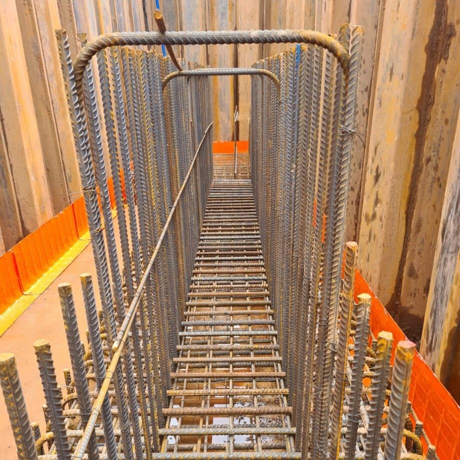 A close-up view of steel rebar arranged in a rectangular cage, likely for structural reinforcement in construction. The bars are placed vertically and horizontally, with wooden and orange safety barriers in the background.