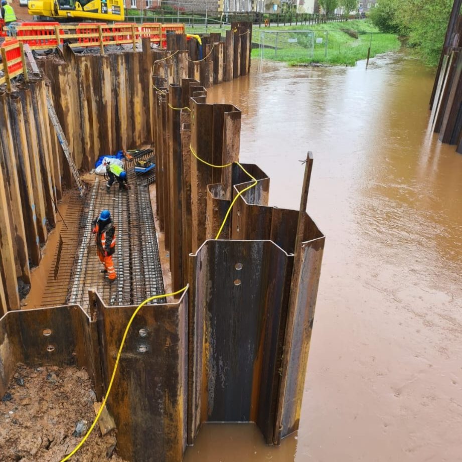 Construction workers in orange protective gear work inside sheet piling on a metal structure along a flooded area. The scene includes muddy water and nearby construction equipment. A yellow cable runs through the structure, highlighting ongoing construction.