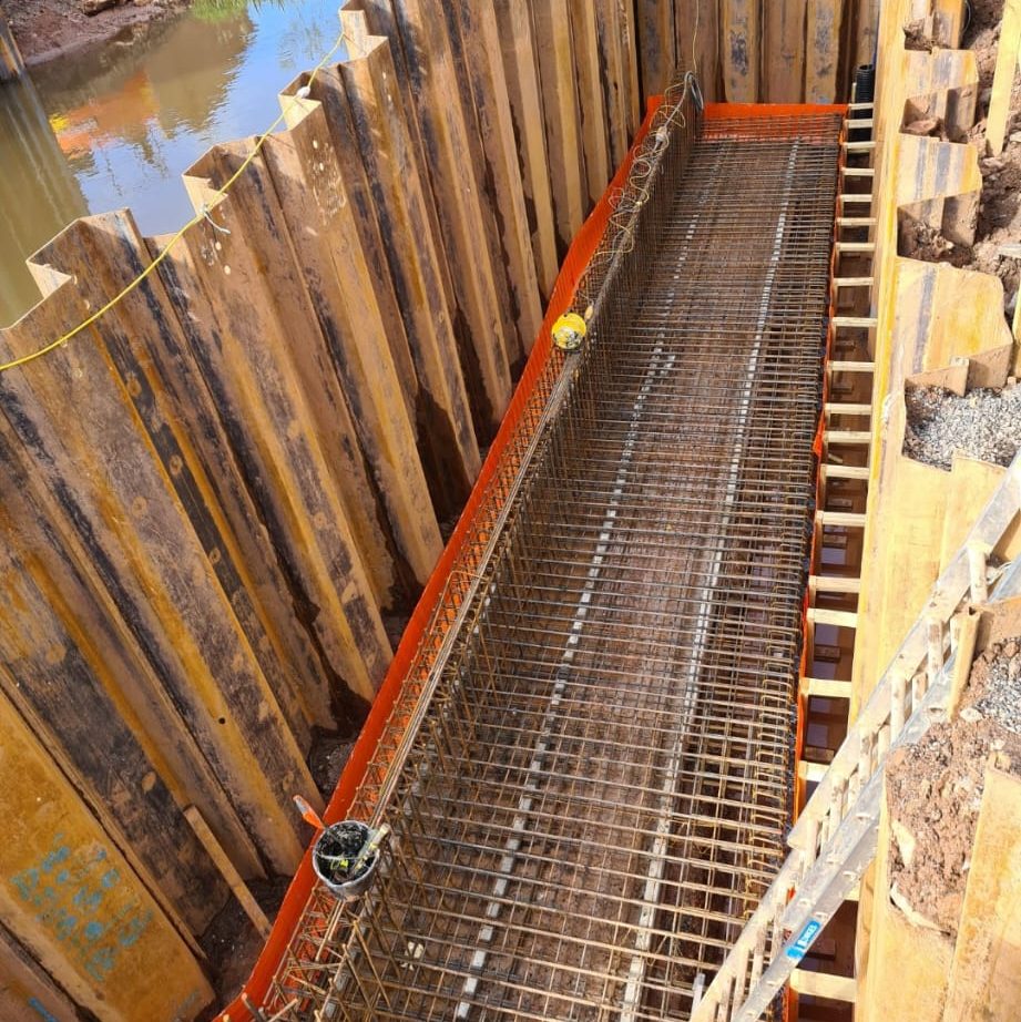 A construction site showing steel rebar arranged within a trench, with wooden supports lining the sides. The trench is prepared for concrete pouring, with water visible in the background.