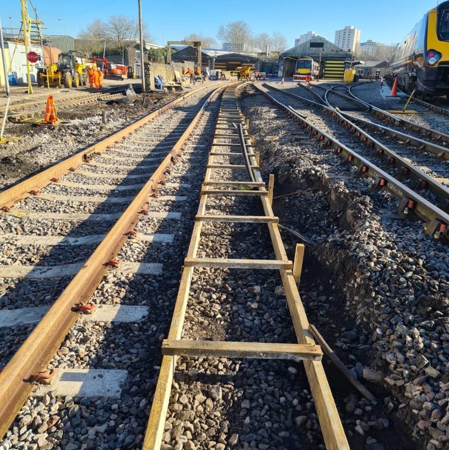 A construction site with train tracks under repair. Wooden planks are laid out on the gravel between the rail tracks. Several trains and construction equipment are visible in the background. Workers and traffic cones are also present on the site.