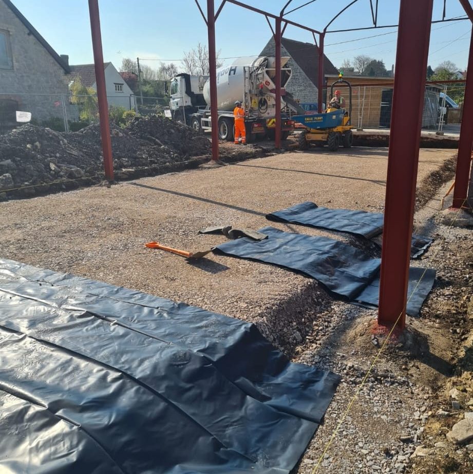 Construction site with a metal frame structure being built. Workers in high-visibility clothing are preparing ground layers with gravel and black sheeting. A concrete mixer truck is in the background, and construction machinery is present on a sunny day.