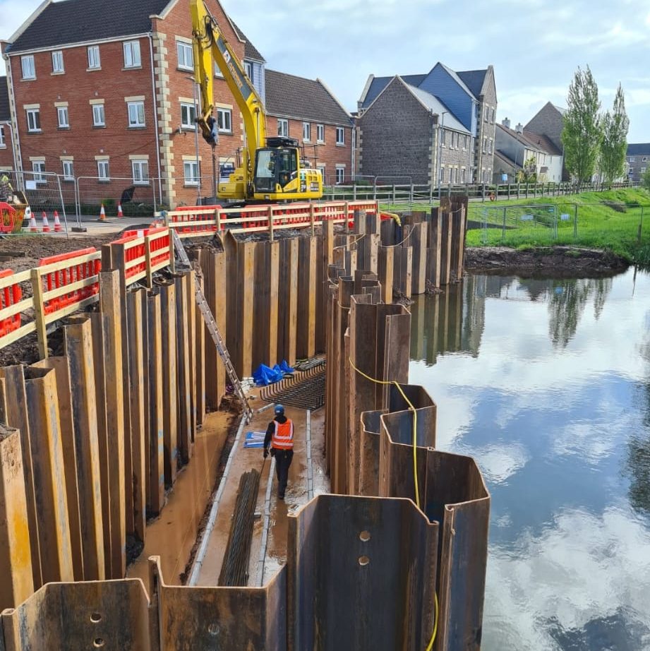 A construction worker in a safety vest stands in a large trench reinforced with steel sheet piles, next to a body of water. An excavator operates nearby, and residential buildings are visible in the background under a cloudy sky.