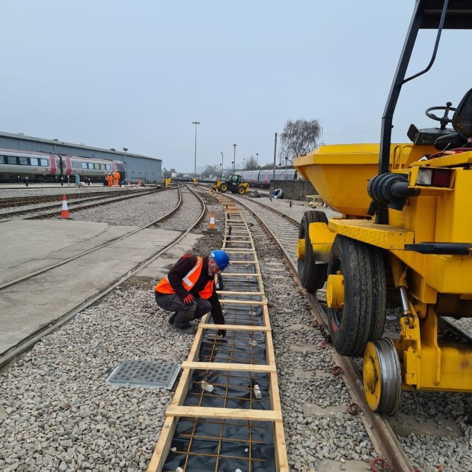 A worker in an orange safety vest and blue helmet kneels on a set of train tracks, inspecting them. Nearby, a yellow rail vehicle is positioned on separate tracks. Multiple train tracks and a distant train are in the background under a cloudy sky.