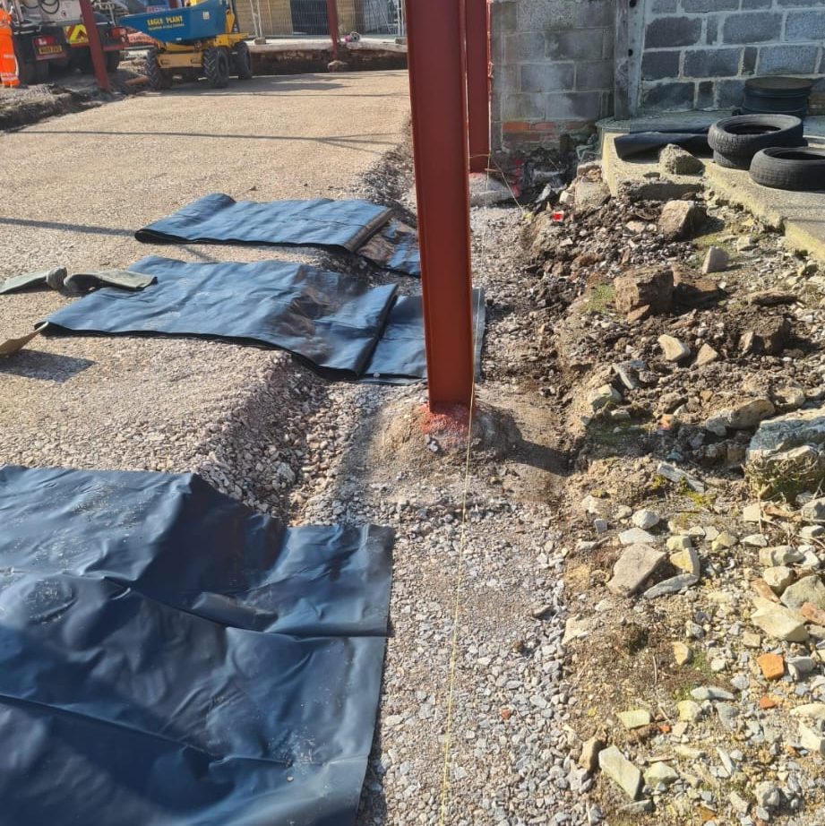 A construction site showing a vertical red steel beam anchored to the ground amidst gravel and rocks. Nearby, black tarps cover sections of the ground. In the background, there are construction materials, equipment, and a partially constructed wall.