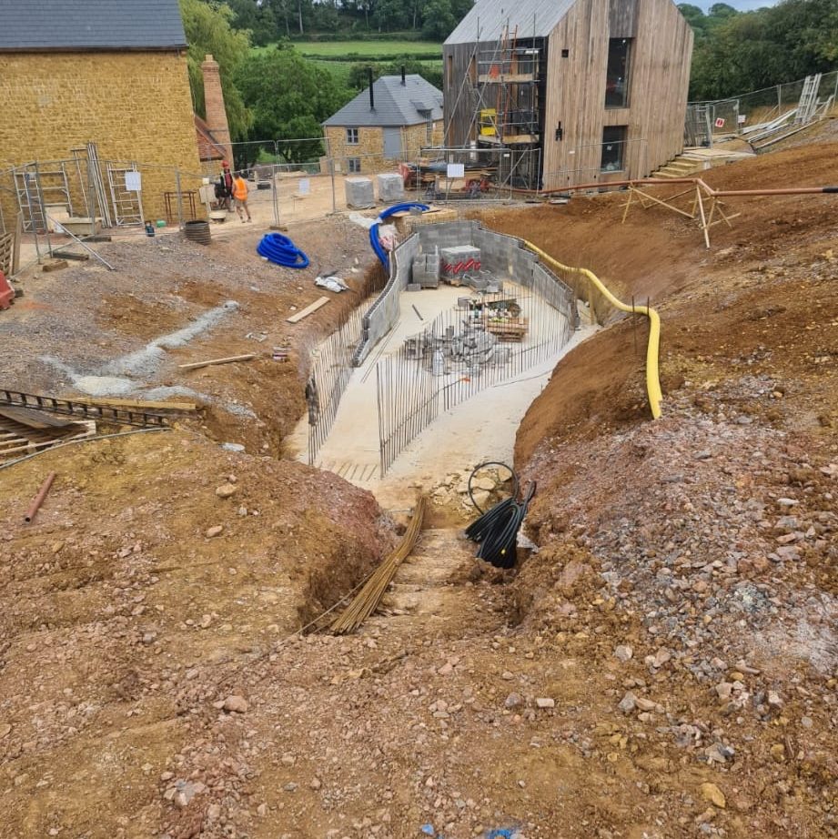 Construction site showing the foundation of a building in progress with exposed earth, concrete forms, rebar, and construction materials. Workers, construction equipment, and partially built structures are visible in the background.