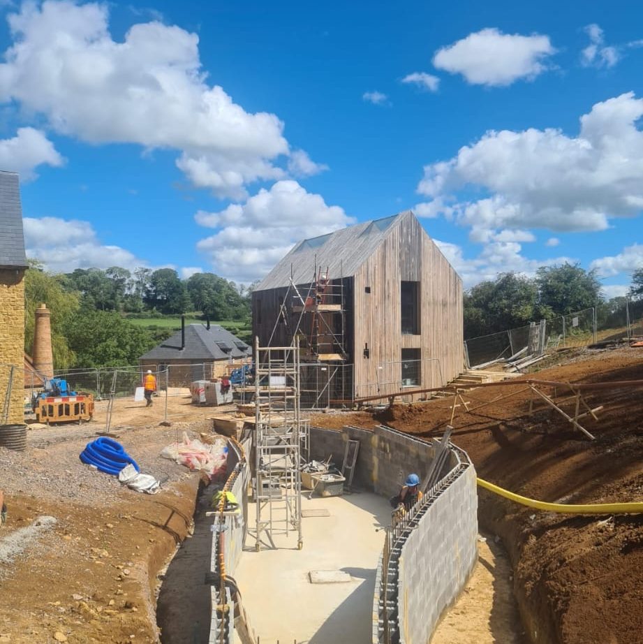 A construction site shows workers building a modern wooden house with large windows. In the foreground, there's a partially built concrete structure, scaffolding, and construction materials. The background features lush green trees and a bright blue sky with scattered clouds.