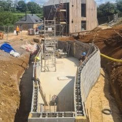 A construction site with workers building a curved foundation wall. Scaffolding is set up inside the foundation, and the area is surrounded by dirt mounds. A modern wooden house and a rural landscape are visible in the background. Construction materials are scattered around.