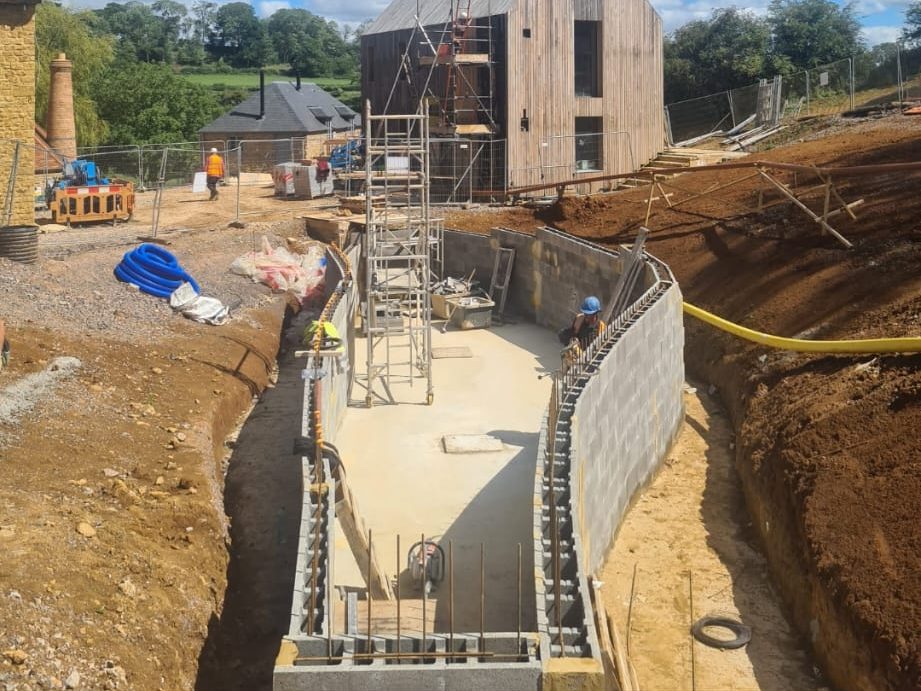 A construction site with workers building a curved foundation wall. Scaffolding is set up inside the foundation, and the area is surrounded by dirt mounds. A modern wooden house and a rural landscape are visible in the background. Construction materials are scattered around.