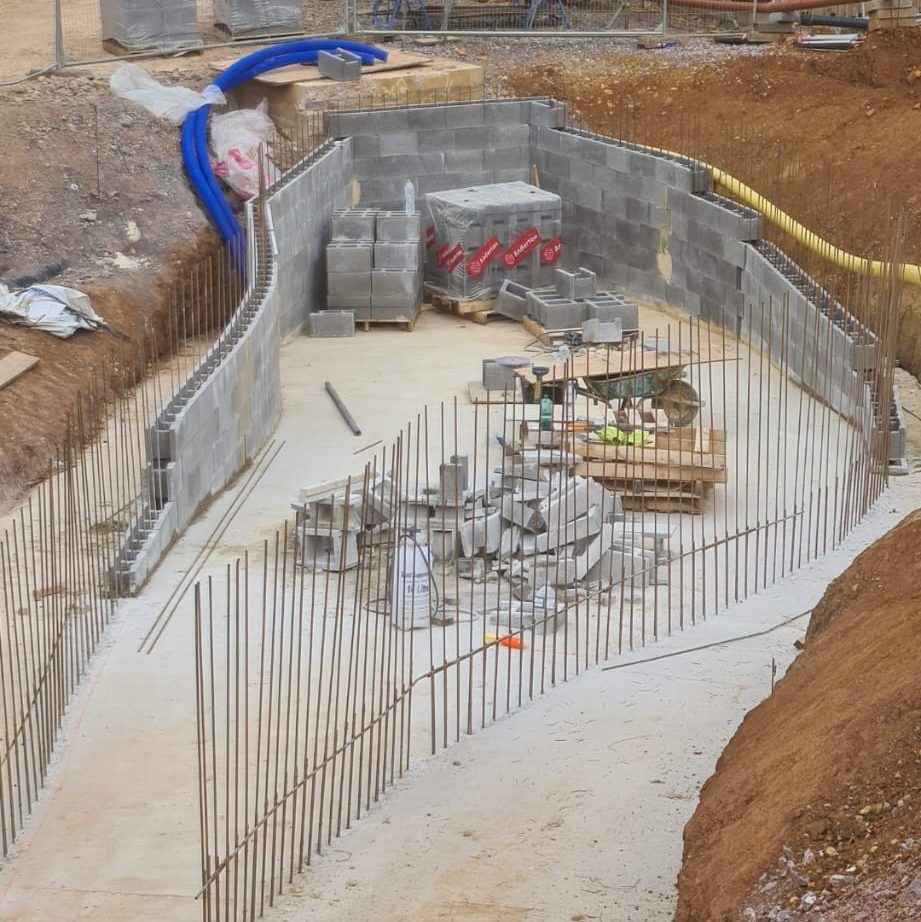 A construction site with a curved concrete foundation and vertical rebar rods. Stacks of concrete blocks, wooden pallets, and construction materials are scattered within the foundation. Surrounding soil and additional construction supplies are visible.