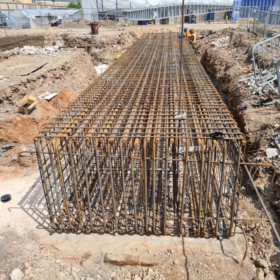 Reinforced steel bars arranged in a rectangular grid within a deep trench at a construction site. The framework is intended for concrete pouring, forming part of the building's foundation. Workers and machinery are visible in the background, surrounded by dirt and construction debris.