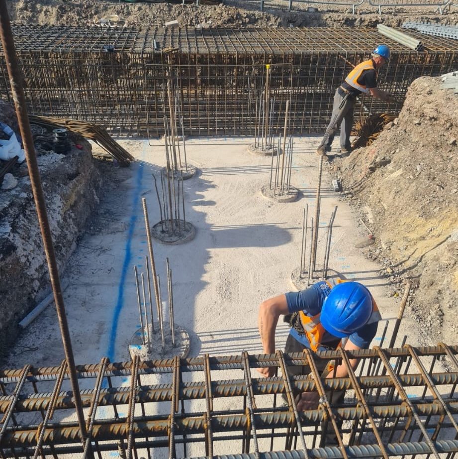 Two construction workers in orange vests and blue helmets are working on a building site, handling rebar for reinforced concrete foundations. The ground is dug out, exposing the rebar structures that form the base of the construction foundation.