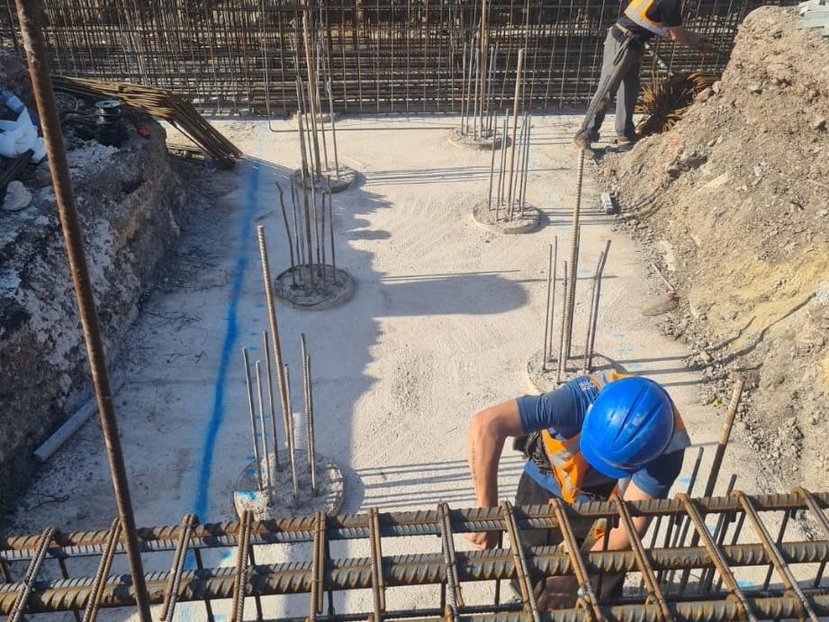 Construction workers at a building site installing vertical rebar for concrete reinforcement. One worker in a blue helmet is placing rebar at the bottom of a foundation, while another worker in a yellow vest is working near the rebar structure.